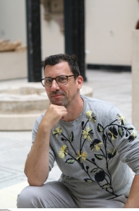Brown-haired white male wearing black glasses and a gray long-sleeve top with yellow & black flowers and gray pants, seated with his right arm resting on his knee and his right hand under his chin 