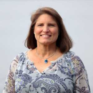 Tammy Euliano author photo showing a smiling white female with brown, shoulder-length hair, wearing a blue-print top and a necklace with a blue stone