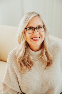 Author JT Ellison: photo of a smiling white woman with long blonde hair, wearing brown eyeglasses, a beige, rolled-neck sweater and a pearl necklace, seated on a beige sofa.