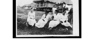 Black and white photograph of several young woman, wearing light-colored, early 20th-century clothing, sitting on a lawn with an old-fashioned school house in the background.