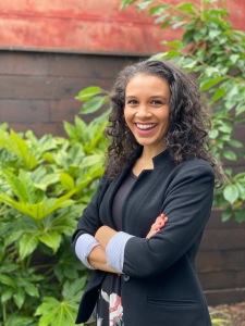 Addie Woolridge author photo - copyright Natasha Beale 2020; smiling Black female with wavy brown shoulder-length hair, wearing a dark blazer and crossed arms standing in front of a dark brick wall with shrubbery in front of the wall