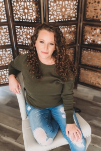Author Marie Still photo: picture of a young, brunette, curly-haired white woman sitting on a light-colored accent chair, wearing denim pants and a dark olive green long-sleeve top