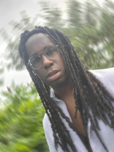 Russell Brooks author photo: head shot photo of a young Black man wearing glasses and a white shirt, with long, brown, locked hair, and blurred greenery in the background