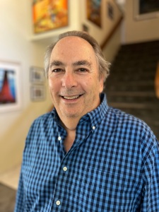 Author Scott Brody: photo of a smiling middlle-aged white male wearing a blue checked shirt standing in front of a staircase