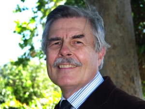 Photo of John G. Bluck: close-up photo of a smiling older white male with salt-and-pepper graying hair and mustache, wearing a striped dress shirt, dark tie, and dark jacket, standing in front of a tree trunk